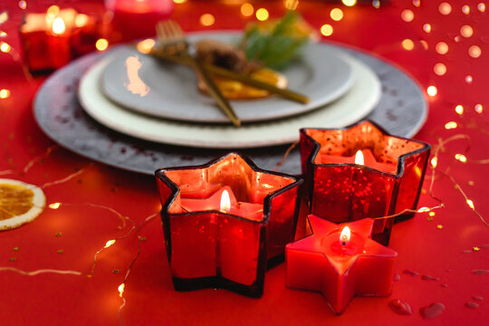 Festive Christmas table setting. Arrangement of candles in form of stars, a plates, golden cutlery, fir branches and cones and sparkles in red and gold tones. Horizontal image.