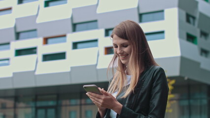 Young Attractive Girl with Long Brunette Hair walking in the Street, Having Mobile Phone in her Hands. Typing Messages and Smiling while sending. Having fashionable clothes.