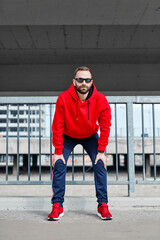 Handsome bearded runner in shape holding hands on knees and taking a break from running in urban...