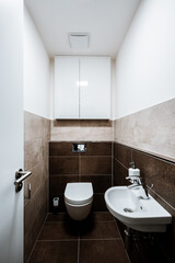 Small modern restroom with all furnishings, including a small sink, cabinet and ceiling fan. Paving and tiling have texture of concrete trowel in shade of brown. The toilet seat is closed.