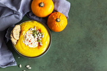 Pumpkin soup served in a bowl with cream, seeds and multigrain crackers. Green concrete background. Top view.