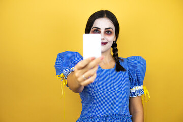 woman wearing a scary doll halloween costume over yellow background smiling and holding white card