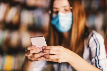 Closeup of young attractive girl sitting in library and using smart phone. Studying during corona pandemic concept.
