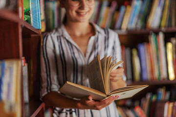 Young smiling attractive college girl leaning on book shelves in library and searching for material for homework.
