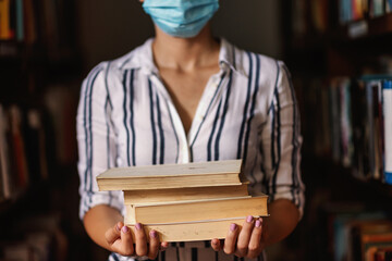 Closeup of girl with face mask standing in library and holding books. Studying during pandemic...