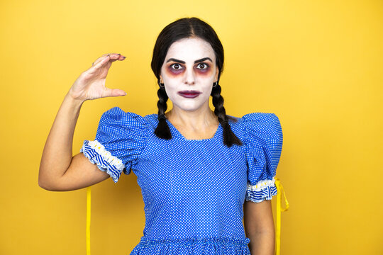 woman wearing a scary doll halloween costume over yellow background smiling and confident gesturing with hand doing small size sign with fingers looking and the camera. Measure concept