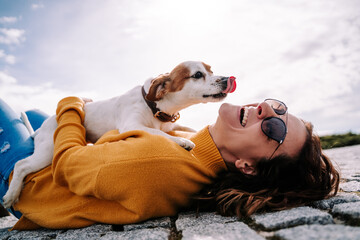 Fototapeta A beautiful woman laughing while her pet is licking her face in a sunny day in the park in Madrid. The dog is on its owner between her hands. Family dog outdoor lifestyle obraz