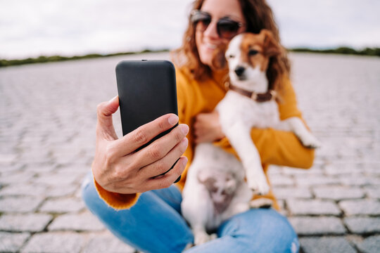 Selective focus. Close-up. A beautiful woman sitting in a park with her cute little dog. She is taking a self-portrait with her dog with the phone. Technology lifestyle with pets