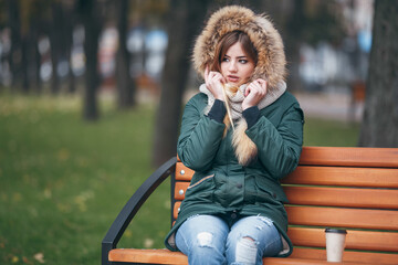Young woman in autumn clothes sits on a bench in a city park. The woman is dressed in a stylish jacket with fur