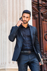 Young East Indian American Businessman traveling, working in New York City, with beard, wearing black suit, carrying laptop computer, walking from old style office doorway, talking on cell phone..