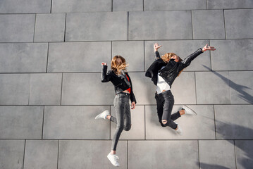 Two beautiful teenage girlfriends jump and having fun near grey concrete wall. Leisure time with best friends.