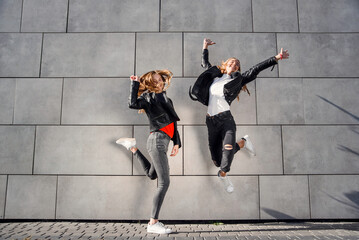 Two beautiful teenage girlfriends jump and having fun near grey concrete wall. Leisure time with best friends.