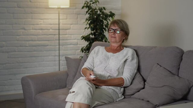 Middle Aged Woman Sitting On The Couch In Her Apartment. She Holds The Remote From The TV And Switches Channels. Light In The Room Changes.