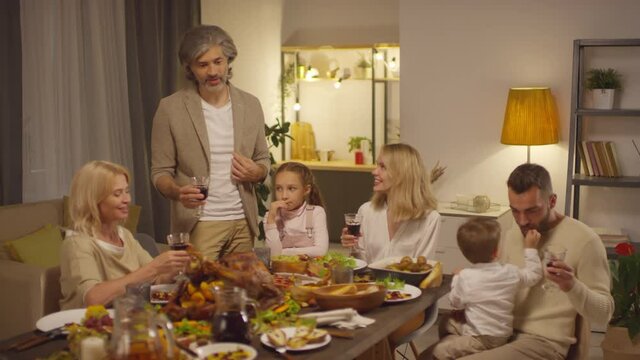 Mature man standing at table proposing toast then clinking glasses with family while celebrating Thanksgiving day