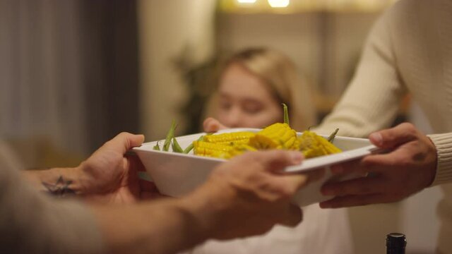 Unrecognizable man passing white ceramic baking dish full of cooked corn to his friend during Thanksgiving day dinner