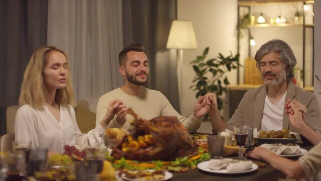 Medium shot of thankful people sitting at dining table holding hands starting celebrating Thanksgiving day with praying