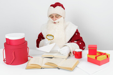 Tired Santa Claus sits at the table and examines letters with a magnifying glass. There are Christmas gifts and books on the table.