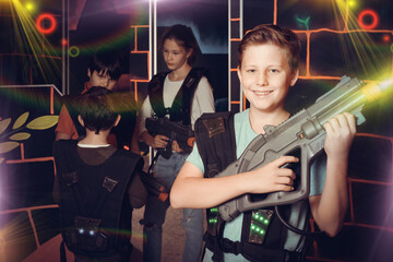 Fototapeta na wymiar Portrait of laughing boy standing with laser gun on dark lasertag arena on background with his team
