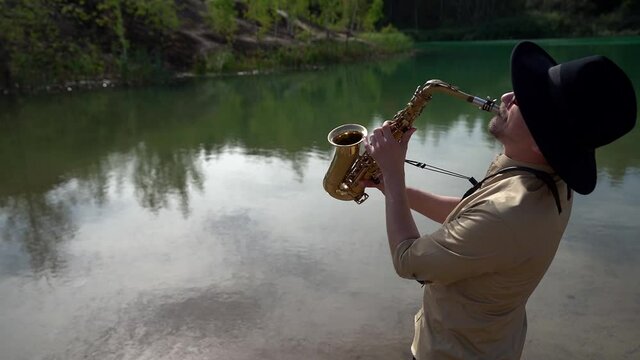jazz musician is playing song by saxophone at nature, man with sax is standing on beach of lake