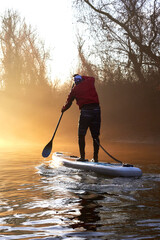 Silhouette of man rowing on SUP (stand up paddle board) at sunrise in a foggy haze in the winter Danube river