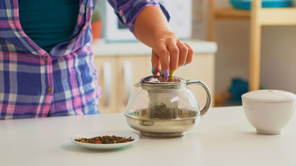 Close up of young woman adding hot water over herbal, tea leaves. Housewife preparing green tea for breakfast using teapot in a modern kitchen sitting near the table holding kettle waiting to infuse.