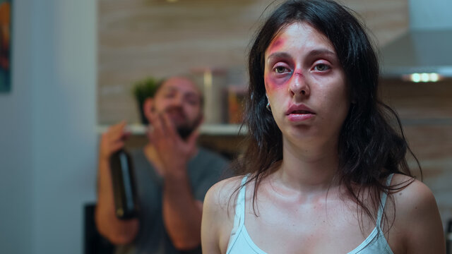Hurt woman and drunk man holding a bottle of wine sitting in the kitchen. Violent aggressive husband abusing injuring terrified helpless, vulnerable, afraid, beaten and panicked wife living in terror.