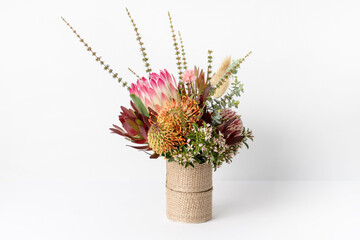 Beautiful Australian native flower arrangement of a red protea flower, orange and red banksias, red leucadendrons, white Eriostemons and eucalyptus leaves, in a rustic vase on white background.