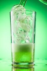pouring green tarragon lemonade into glass with pieces of pure real ice on green gradient background