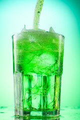 pouring green tarragon lemonade into glass with pieces of pure real ice on green gradient background
