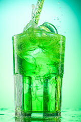 pouring green tarragon lemonade into glass with pieces of pure real ice on green gradient background