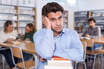 Portrait of weary upset adult man with stack of books in hands in public library..