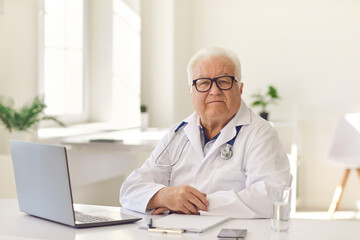 Serious senior doctor sitting at desk with laptop in modern hospital office looking at camera