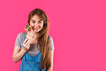 pretty caucasian blonde girl in denim dress with lollipop in her hand. smile and look straight. close up. isolated on pink background.