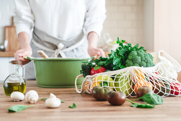 Woman cooking with fresh organic food in the background. Fruits and vegetables in cotton reusable net mesh bag on wooden table in modern bright kitchen