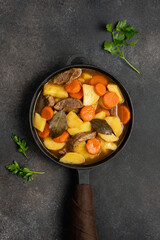 Stew with carrots and potatoes in cast iron skillet on dark brown concrete background. Seasonal home cooking
