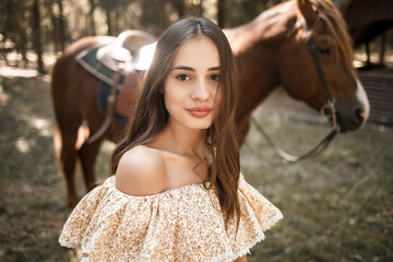 A beautiful young girl dressed in a dress stands near a horse in the forest