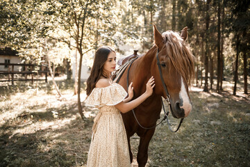 Fototapeta premium A beautiful young girl dressed in a dress stands near a horse in the forest