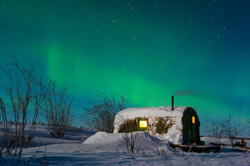 Night arctic landscape. Snow-covered hunting lodge in the tundra. Northern lights and stars in the night sky. Northern nature of Chukotka and polar Siberia. Travel to the extreme North of Russia.