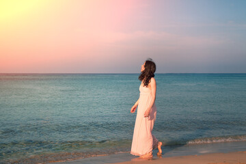 Fototapeta na wymiar A young happy woman in a long dress walks on the beach at sunrise. Summertime