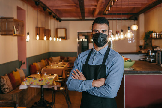 Serious owner crossing arms on chest during Coronavirus pandemic. African american waiter wearing a face mask, standing in the dining room of a restaurant and looking at camera with determination.