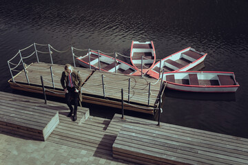 An attractive young guy in a khaki military cloak stands on a boat pier (river boat station). Top view of four empty boats.