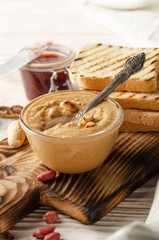 Low angle view at glass bowl with peanut butter on white wooden table with toasts and jam aside. Healthy eating concept