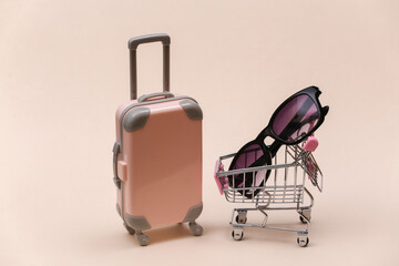 Travel and shopping concept. Mini plastic travel suitcase and supermarket trolley with sunglasses on beige background.