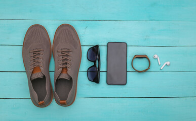 Men's shoes and gadgets (smartphone, smart bracelet, headphones), sunglasses on a blue wooden background. Top view