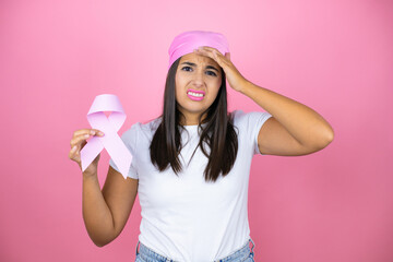 Young beautiful woman wearing pink headscarf holding brest cancer ribbon over isolated pink background stressed and frustrated with hand on head, surprised and angry face