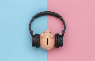 Piggy bank listens stereo headphones on blue-pink pastel background. Top view