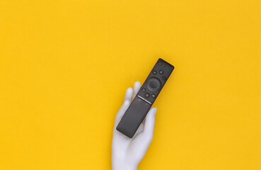 White mannequin hand holds tv remote on yellow background. Top view
