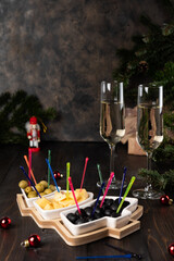 Christmas tree plate with green and black olives, cheese. Glasses of champagne, nutcracker and fir tree branches on old wooden background, dark image in rustic style, selective focus