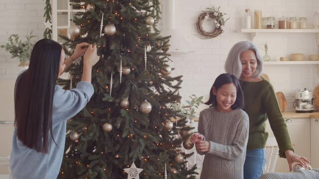 Happy Asian girl decorating Christmas tree with mother and grandmother while preparing home for celebration