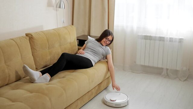 Smiling young brunette woman with glasses lying on sofa touches white round automatic robot vacuum cleaner hoovering floor in living room